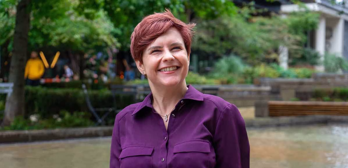 Image of Sassy Wyatt wearing a purple blouse and smiling at the camera. She is standing in front of a pond in a city centre.