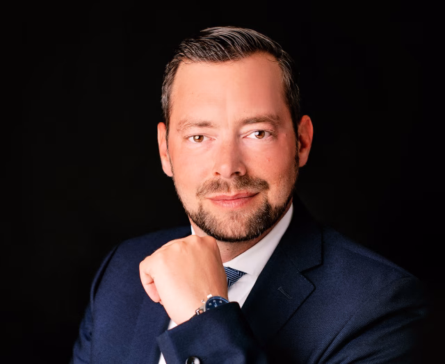Image shows Oliver Kuhn seated against a dark background. He is smiling at the camera.