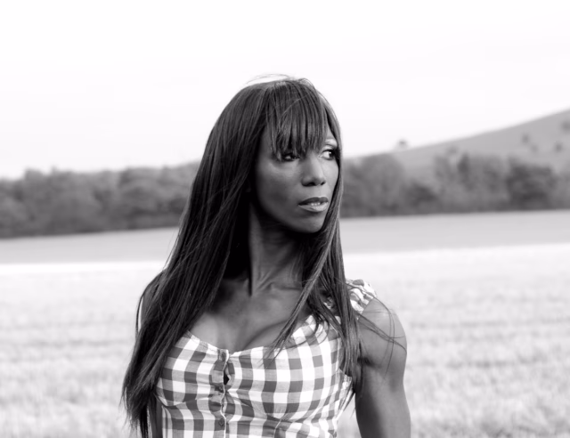 Image shows Angel Sinclair looking over her left shoulder. She is stood against the backdrop of the countryside